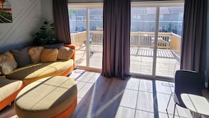Ein Zimmer mit einer Couch, einem Sessel, einer Deckenleuchte und Blick ins Freie.