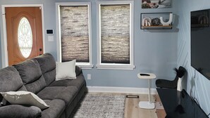 Ein Wohnzimmer mit grauem Ecksofa, einem kleinen Teppich, einem Fernseher und zwei Fenstern mit Jalousien.