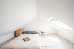 A minimalist bedroom with a sloped ceiling, a bed with rolled towels, a small wooden nightstand, a potted plant, and a skylight.