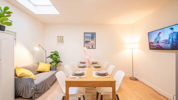 Un coin repas avec une table en bois dressée pour six personnes, des chaises blanches, un canapé gris avec des coussins jaunes, une plante en pot et un téléviseur fixé au mur.