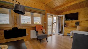 Interior de una cabaña de madera con sofá, una silla, una mesa, televisor y ventanas amplias.