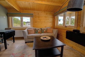 A cozy room with a wooden ceiling, a sofa with colorful cushions, a table with a remote control, and a window with a view of greenery.