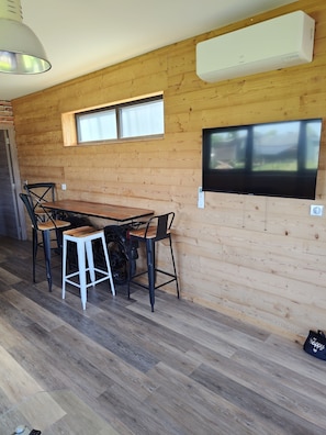 Ein Zimmer mit Holzwänden, einem Esstisch, Stühlen, einem an der Wand befestigten Fernseher und einer Klimaanlage.
