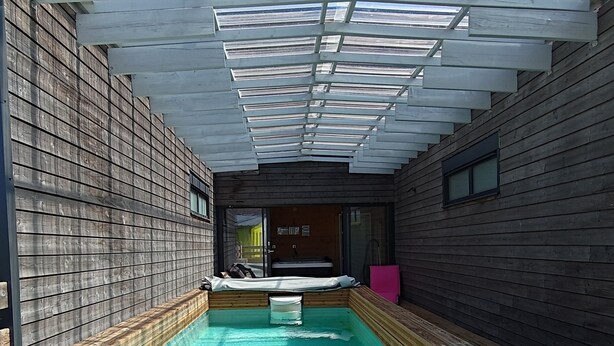 Una piscina con cubierta de madera, entrada por puerta de cristal y vista del cielo a través de una estructura en el techo.