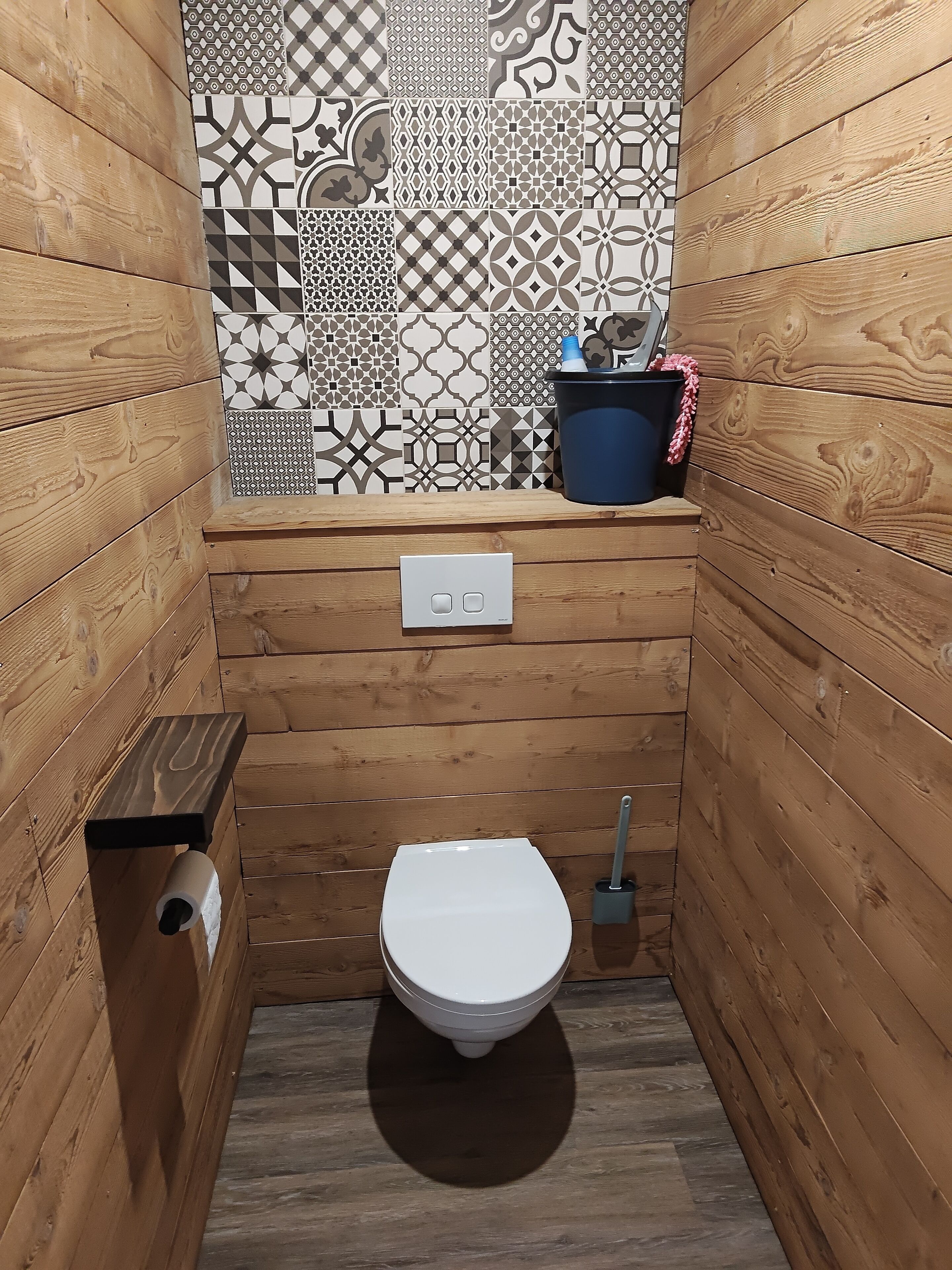 A small, wooden-paneled bathroom with a white toilet, a wall-mounted flush handle, and a patterned tile backsplash.