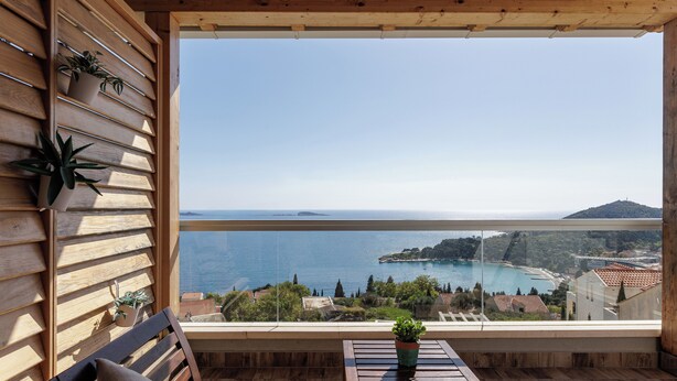 Un balcón con listones de madera, una banca de madera, una mesita con una planta en maceta y vista al mar y al paisaje distante.