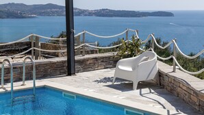 Una piscina con sillones de descanso y una mesa, rodeada por un muro de piedra y una barrera de cuerda, con vista al mar y colinas distantes.