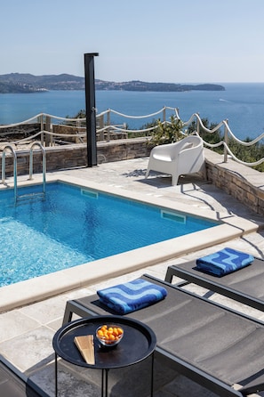 Ein Swimmingpool mit Liegestühlen und einem Tisch, umgeben von einer Steinmauer und einem Seilabfangnetz, mit Blick auf das Meer und ferne Hügel.