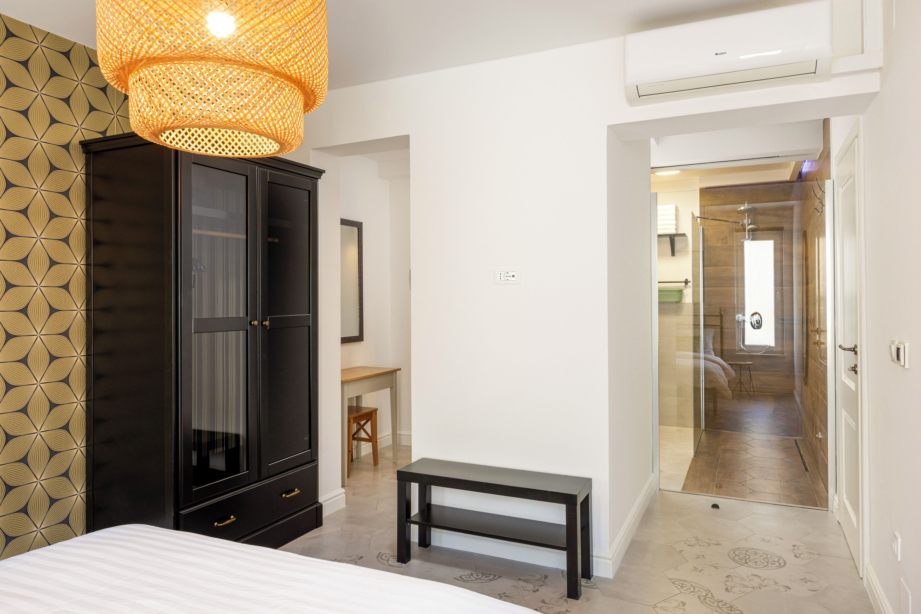 Un dormitorio con cama, armario, banco y baño visible a través de una puerta de cristal.