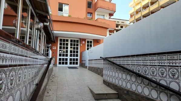 Un étroit chemin menant à l’entrée d’un bâtiment, avec un mur recouvert de carreaux d’un côté et une rambarde métallique de l’autre.