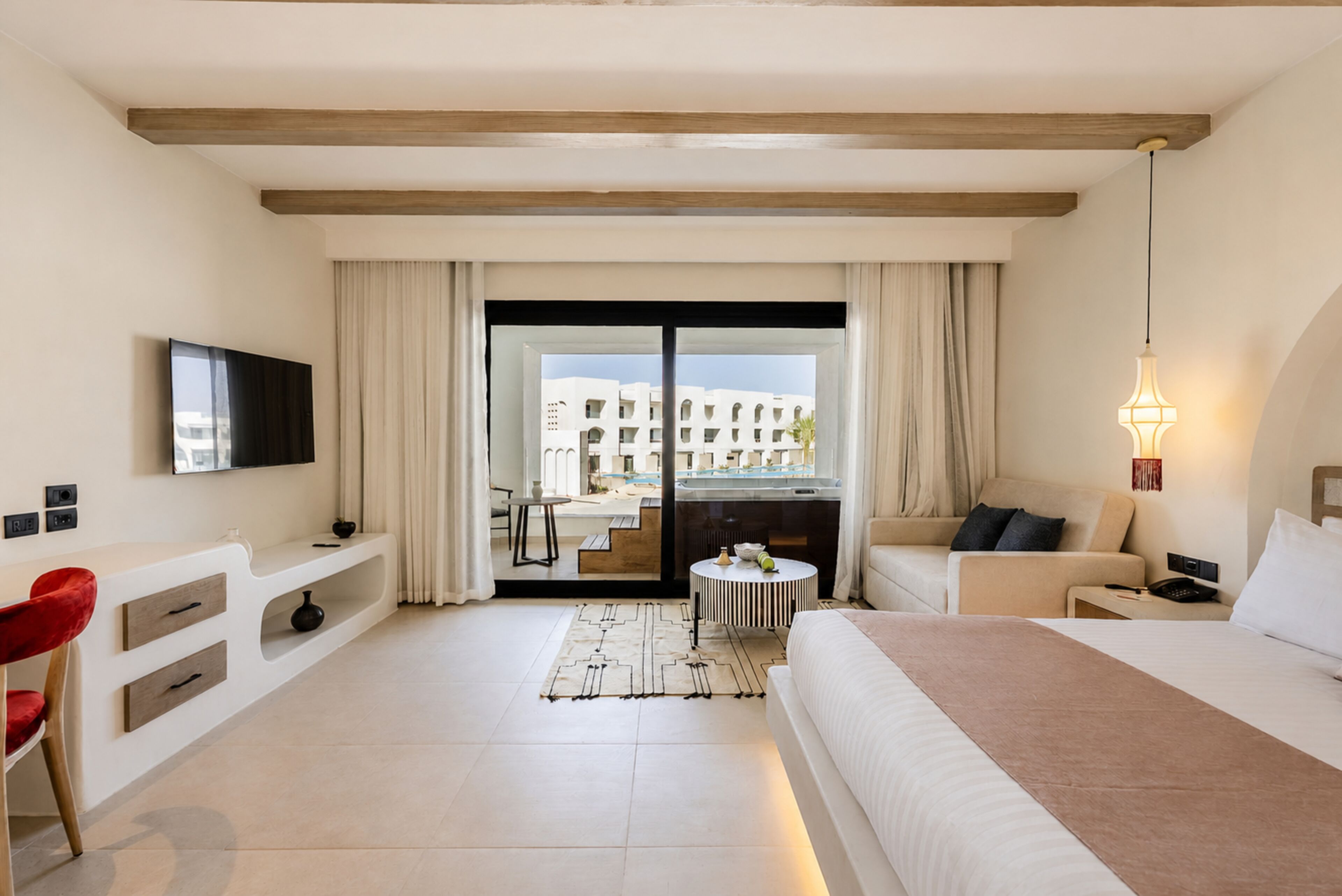 A hotel room with a bed, a desk, a chair, a TV, a balcony with a view of buildings, and a lamp.