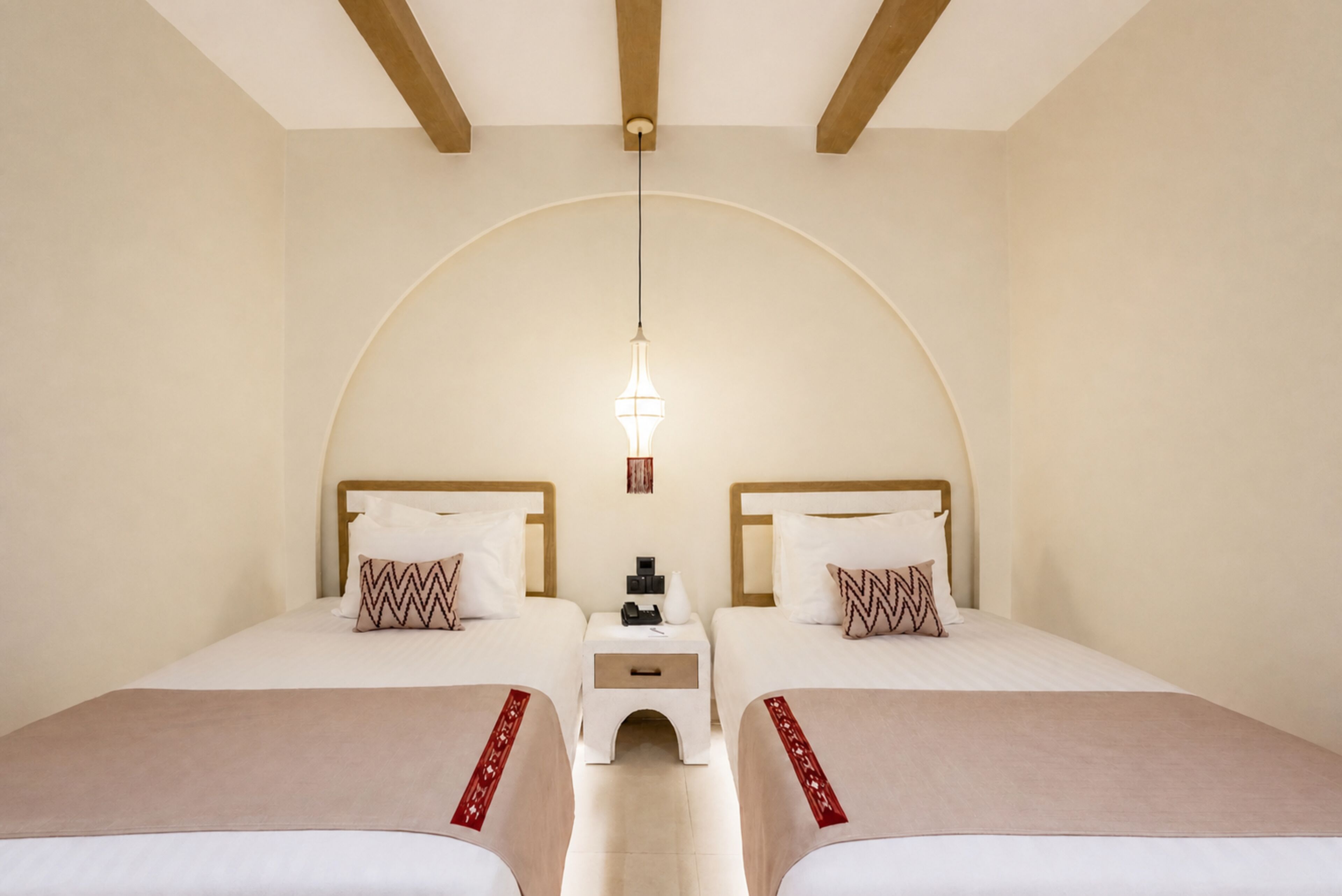Habitación de hotel con dos camas, una mesita de noche pequeña con un teléfono y una lámpara colgante.