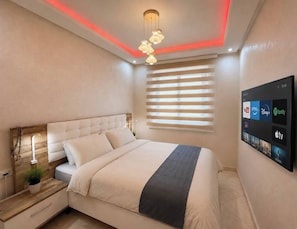 A modern bedroom with a bed, bedside tables, a wall-mounted TV, and a ceiling light fixture. - Open-Plan 2BR Agadir Apartment | WiFi & Parking (Agadir)