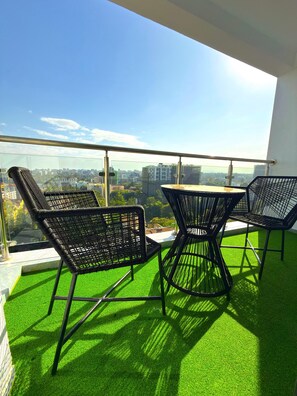 Un balcón con césped artificial verde, sillas de mimbre negro y barandilla de vidrio con vistas a un paisaje urbano.