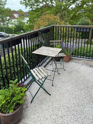 En balkon med et træbord og stole, omgivet af et sort metalgelænder. Der er plantekasser med planter på balkonen.