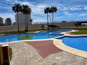 Uma piscina com caminho de pedra e tijolo, palmeiras e uma ponte ao fundo.