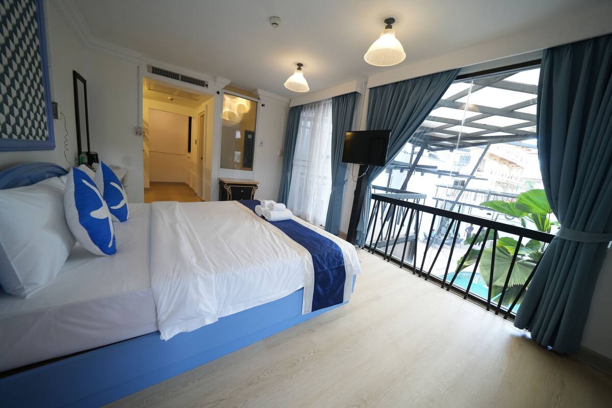 A bedroom with a bed, a TV, a balcony with a railing, and a view of buildings and greenery.