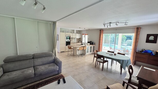 Uma sala de estar com um sofá cinzento, uma área de refeições com mesa e cadeiras, uma cozinha com armários e eletrodomésticos, e uma grande janela com cortinas.