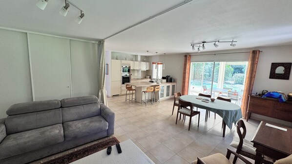 Ein Wohnzimmer mit grauem Sofa, eine Essecke mit Tisch und Stühlen, eine Küche mit Schränken und Geräten sowie ein großes Fenster mit Vorhängen.