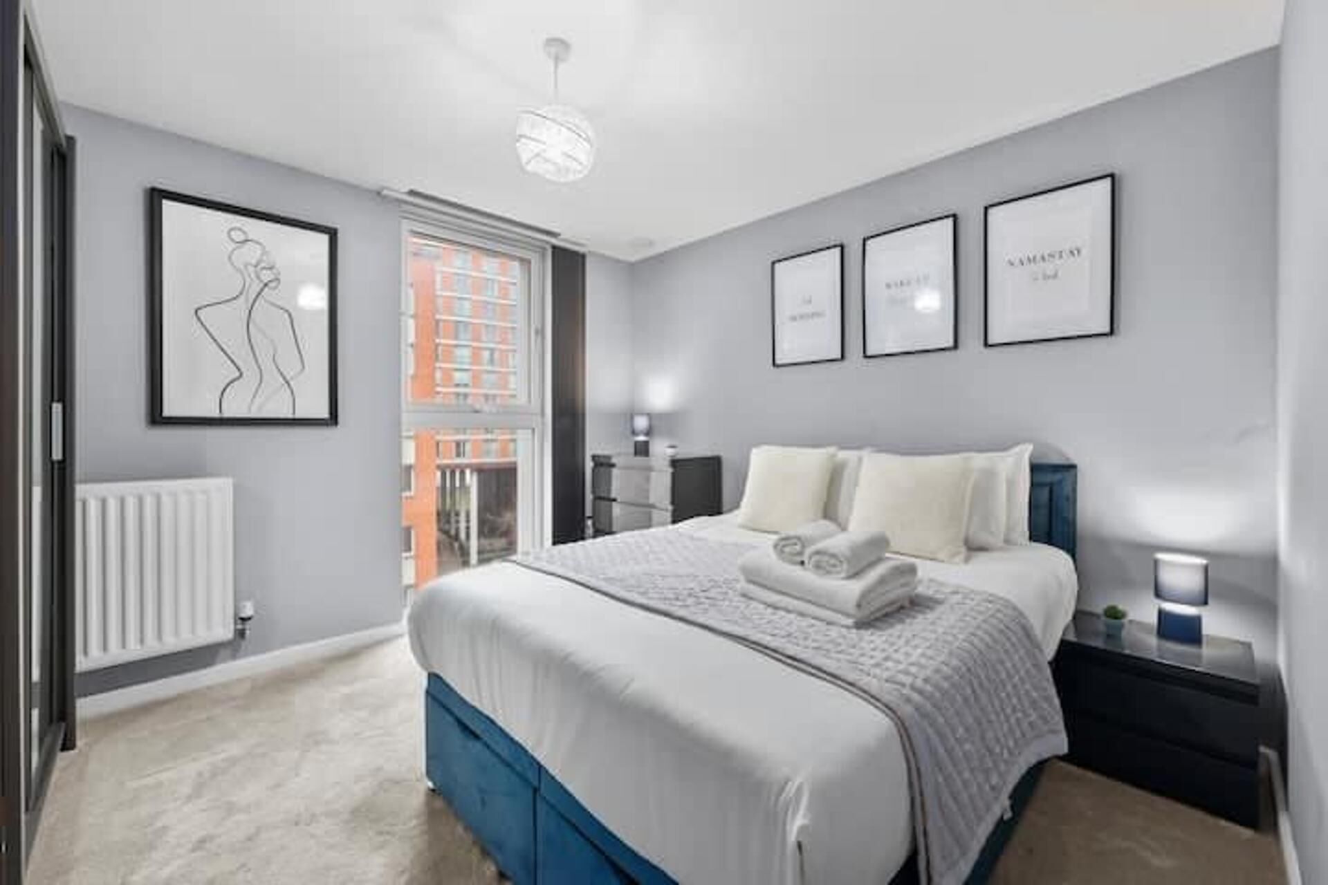 A modern bedroom with a large bed, a radiator, a window, and framed artwork on the wall.