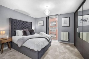 A modern bedroom with a tufted headboard, a large bed, a nightstand, a mirror, and a framed quote on the wall. - 1 Bed Apartment Sleeps 3 With Balcony (London)