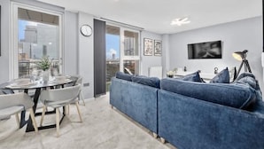 Een moderne woonkamer met een blauwe bank, een eettafel en een stadszicht door grote ramen.