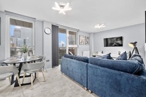 A modern living room with a blue sofa, a dining table set, and a city view through large windows. - 1 Bed Apartment Sleeps 3 With Balcony (London)