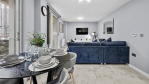 Een moderne woonkamer met een blauwe bank, een eettafel voor vier personen, een flatscreen televisie aan de muur en een groot raam.