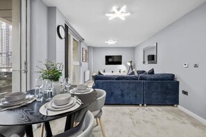A modern living room with a blue sofa, a dining table set for four, a flat-screen TV mounted on the wall, and a large window. - 1 Bed Apartment Sleeps 3 With Balcony (London)