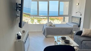 Habitación de hotel con cama, sofá, televisión montada en la pared, una mesa con una bandeja de tazas y gofres, y vista al mar a través de amplias ventanas.