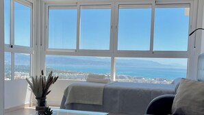 Un moderno salón con un sofá, una mesa de centro de cristal y vistas al mar y al paisaje urbano a través de amplios ventanales.