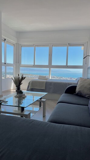 Ein modernes Wohnzimmer mit einer Couch, einem Glastisch und Meer- und Stadtblick durch große Fenster.
