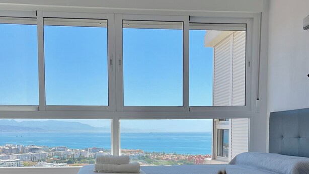 Un dormitorio con una ventana grande, una cama, una mesita de noche con un florero y vistas al mar.