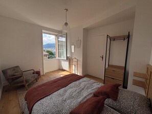 A bedroom with a large bed, a chair, a wardrobe, and a window with a view of buildings. - Los Arcos | Two renovated houses for 15 people. 4-legged friends too! (Annonay)