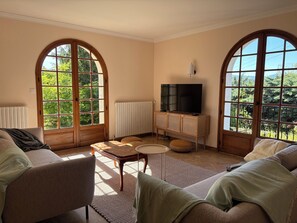 A living room with a sofa, armchair, and ottoman, a television on a wooden cabinet, and large arched windows with a view of greenery. - Los Arcos | Two renovated houses for 15 people. 4-legged friends too! (Annonay)