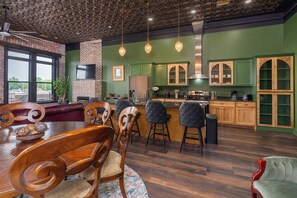 Een eethoek met houten stoelen en een tafel, een bar met krukken, een keuken met groene kastjes, en een bakstenen muur.