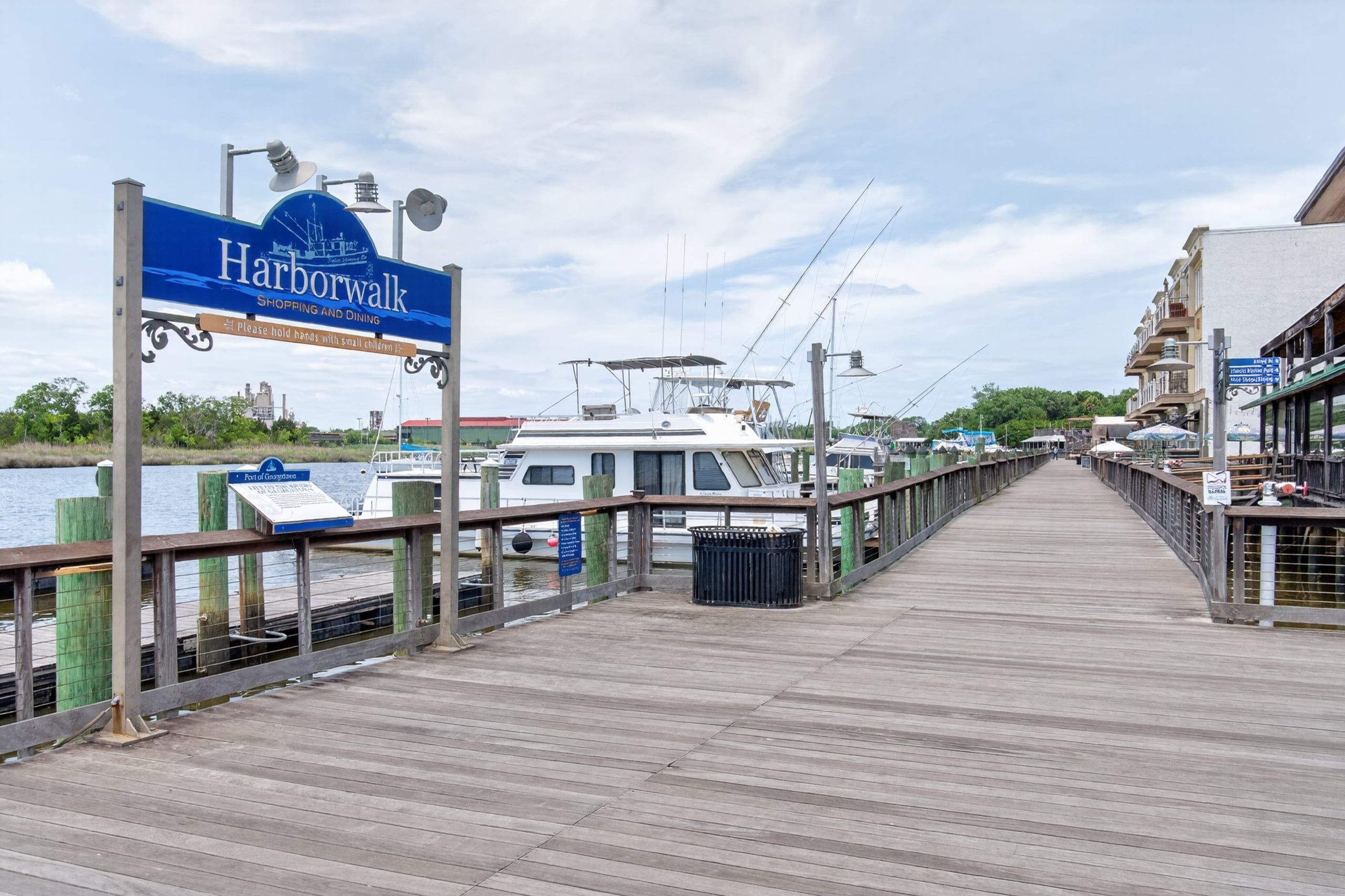 A wooden boardwalk leading to a marina with boats docked, a sign for Harborwalk, and a view of buildings and greenery.