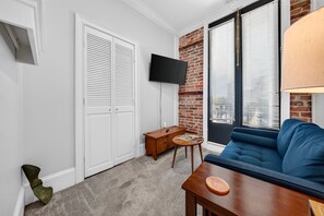 A living room with a blue sofa, a wooden coffee table, a TV mounted on the wall, and a brick accent wall. - Harbor View Suite B in Historic Georgetown (Georgetown)