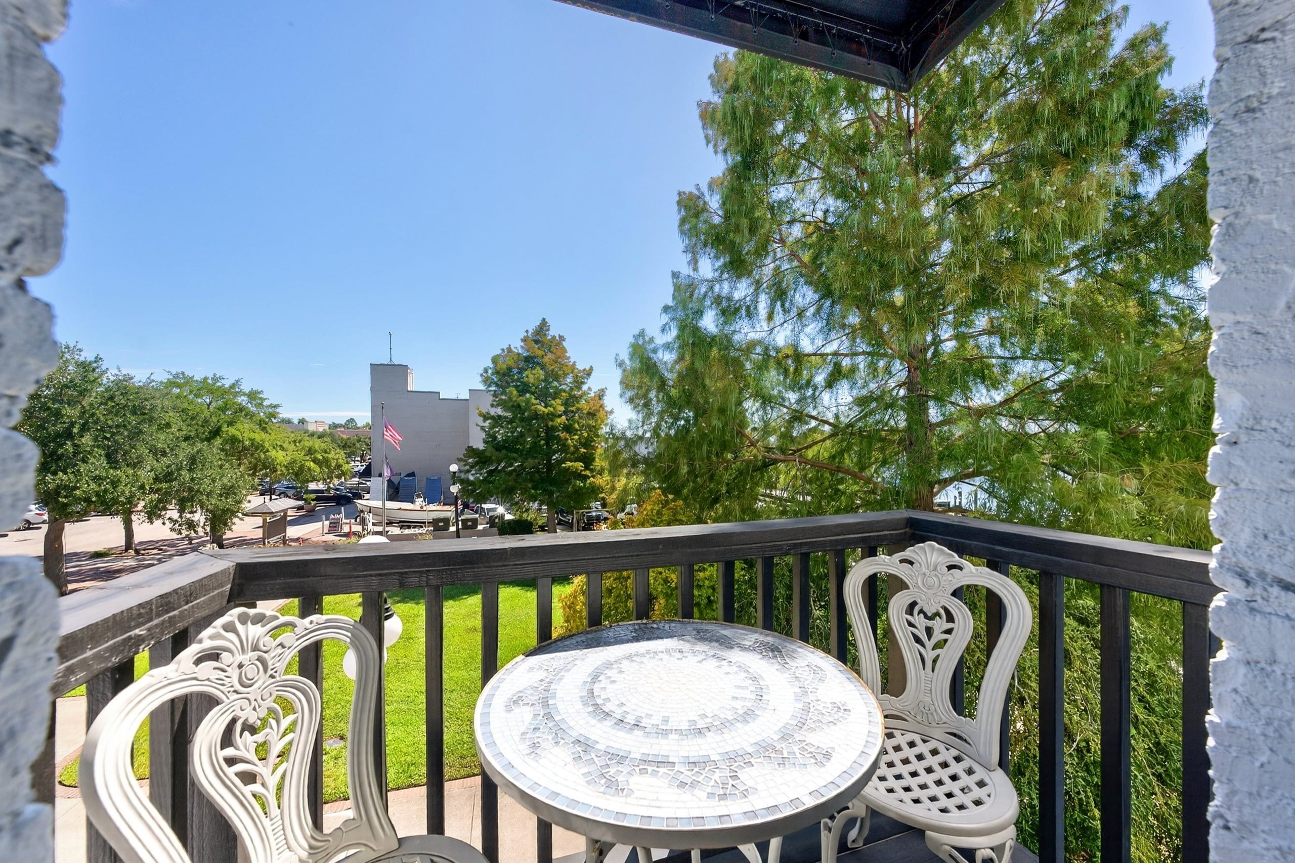 A balcony with a round table and two chairs, overlooking a parking area and a building with a flagpole.