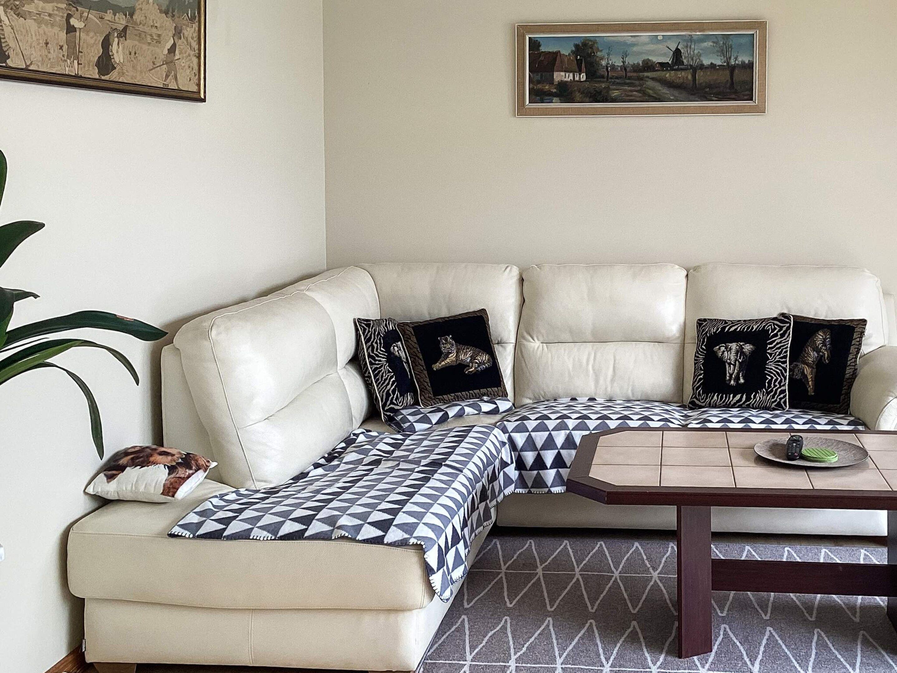Una sala de estar con esquina, un sofá de cuero color beige, almohadones decorativos estampados, una pequeña mesa de centro de madera y un cuadro con un paisaje en la pared.