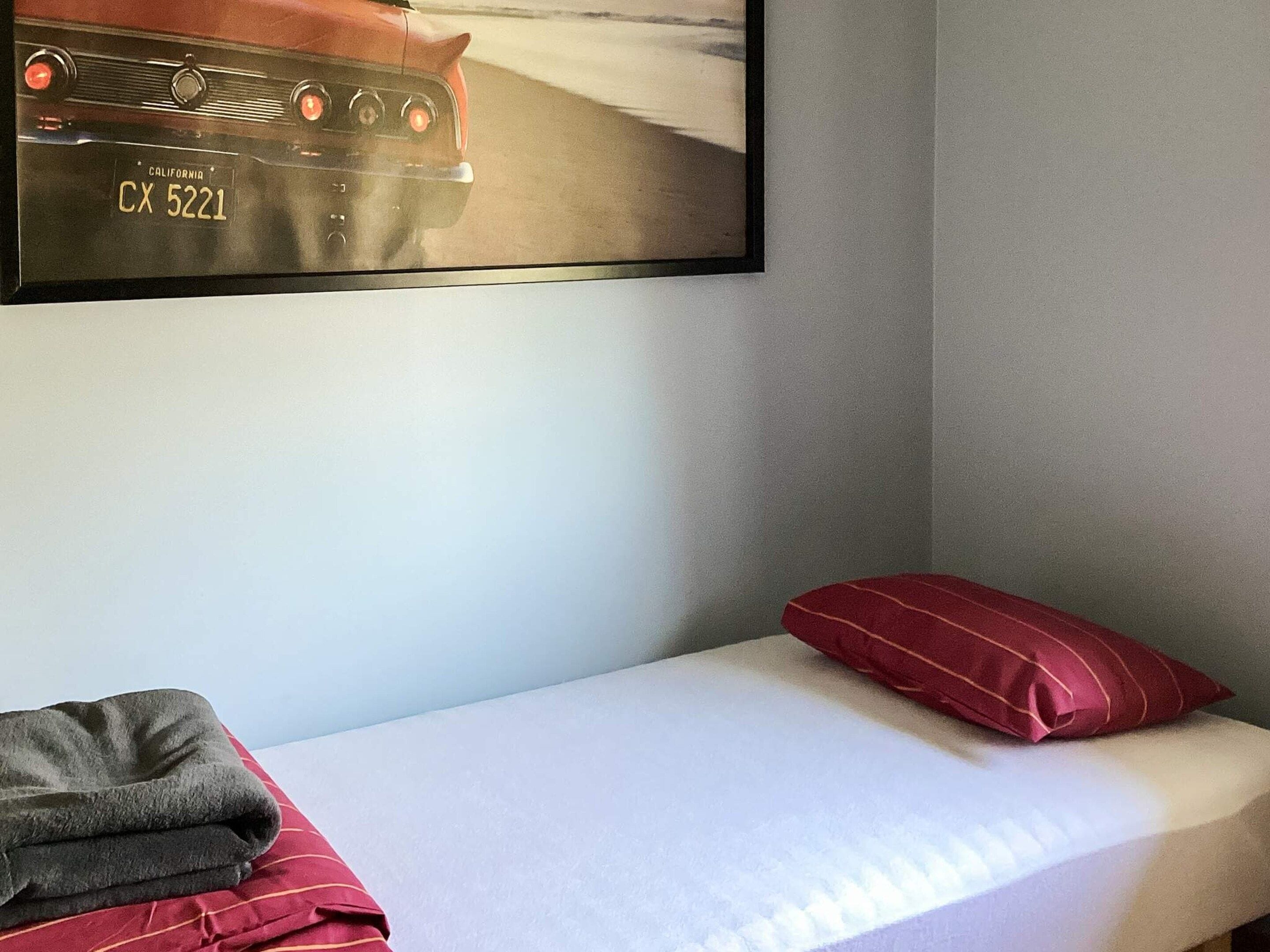 A single bed with a red pillow and a folded blanket, a framed picture of a car on the wall, and a grey wall.