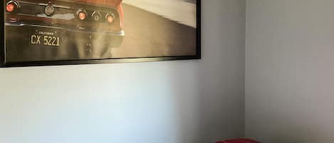 A single bed with a red pillow and a folded blanket, a framed picture of a car on the wall, and a grey wall.