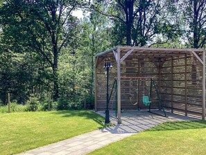 En pergola av trä med en gunga placerad på en gräsyta, omgiven av träd.