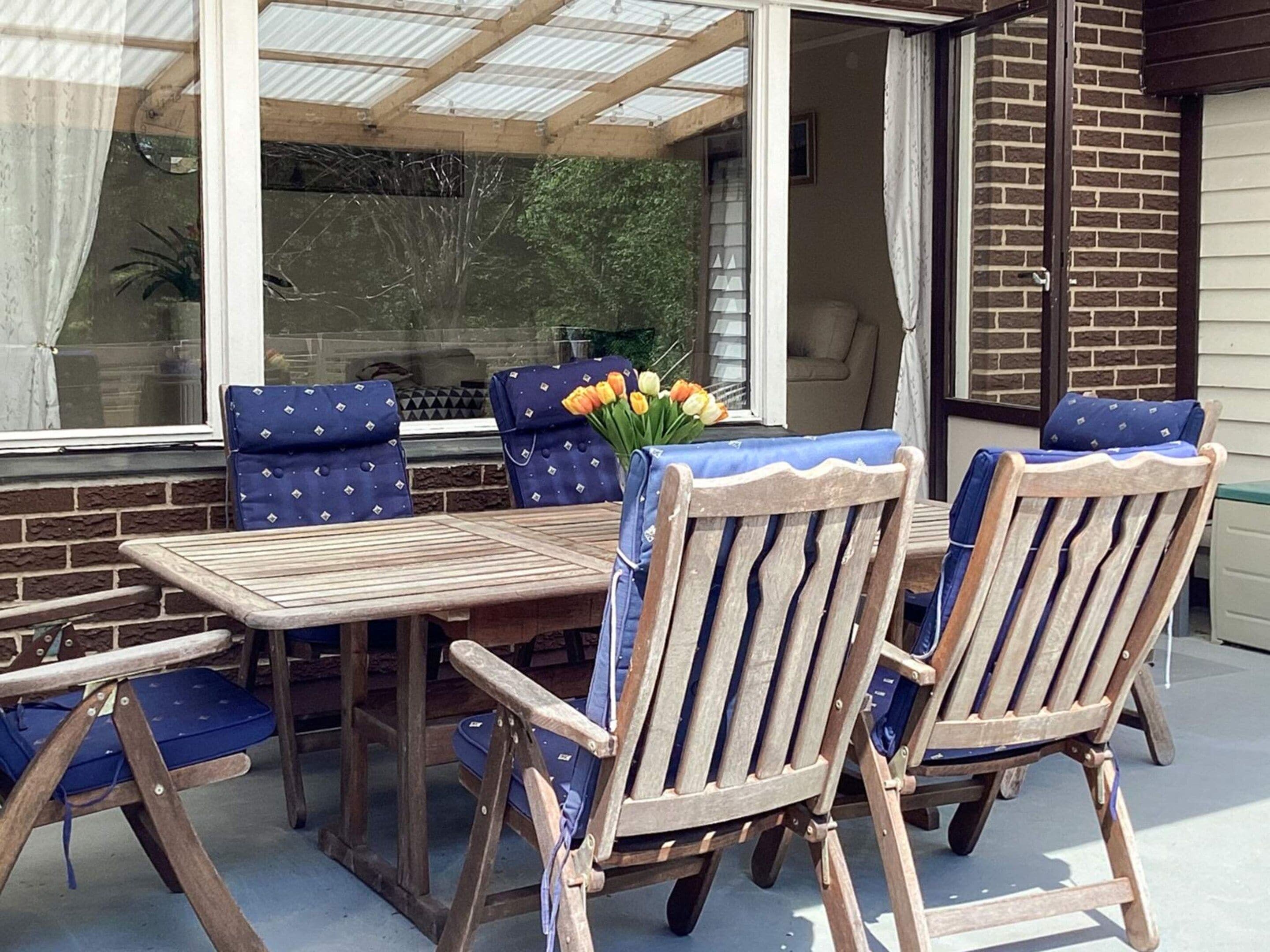 Eine Terrasse mit Holztisch und -stühlen, einer Glaskonstruktion und einem Fenster mit Vorhängen.