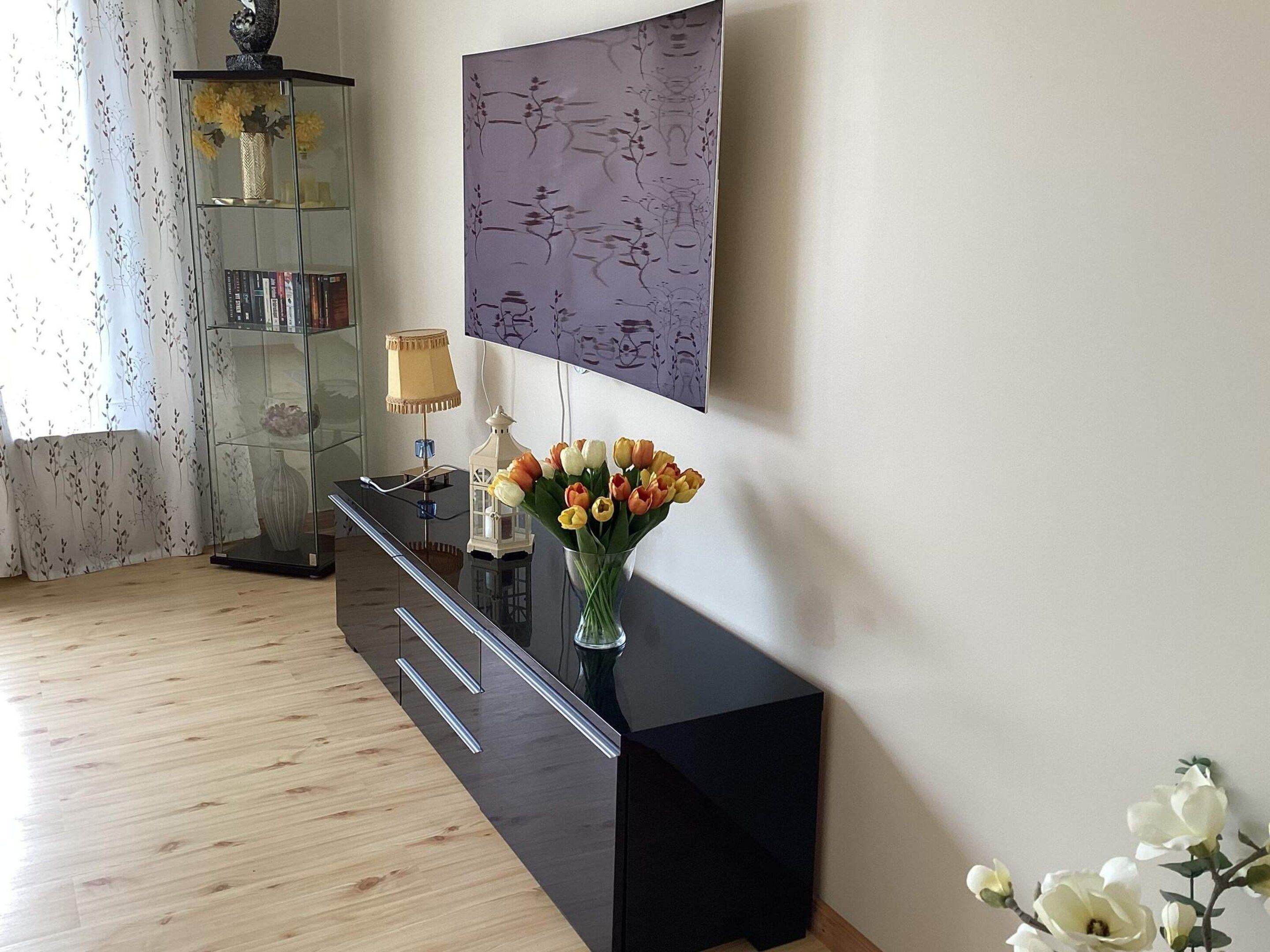 A room with a black console table, a vase with flowers, a wall-mounted artwork, and a glass cabinet with decorative items.