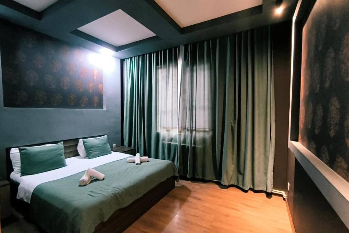 A hotel room with a bed, curtains, a painting on the wall, and a ceiling light.