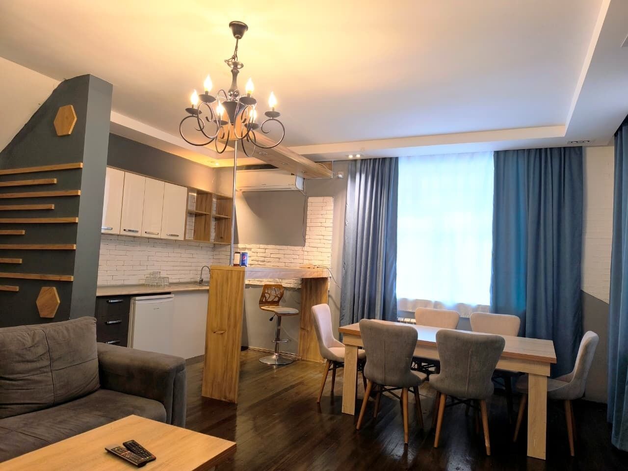 A modern living room with a sofa, dining table, chairs, a chandelier, and a kitchen area with cabinets and a countertop.