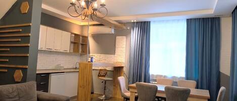 A modern living room with a sofa, dining table, chairs, a chandelier, and a kitchen area with cabinets and a countertop.
