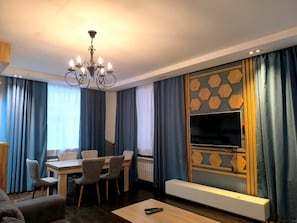 A modern living room with a dining area, a chandelier, a flat-screen TV mounted on a geometric wall, and a sofa. - Fortuna Boutique Hotel & Restaurant (Gabala)