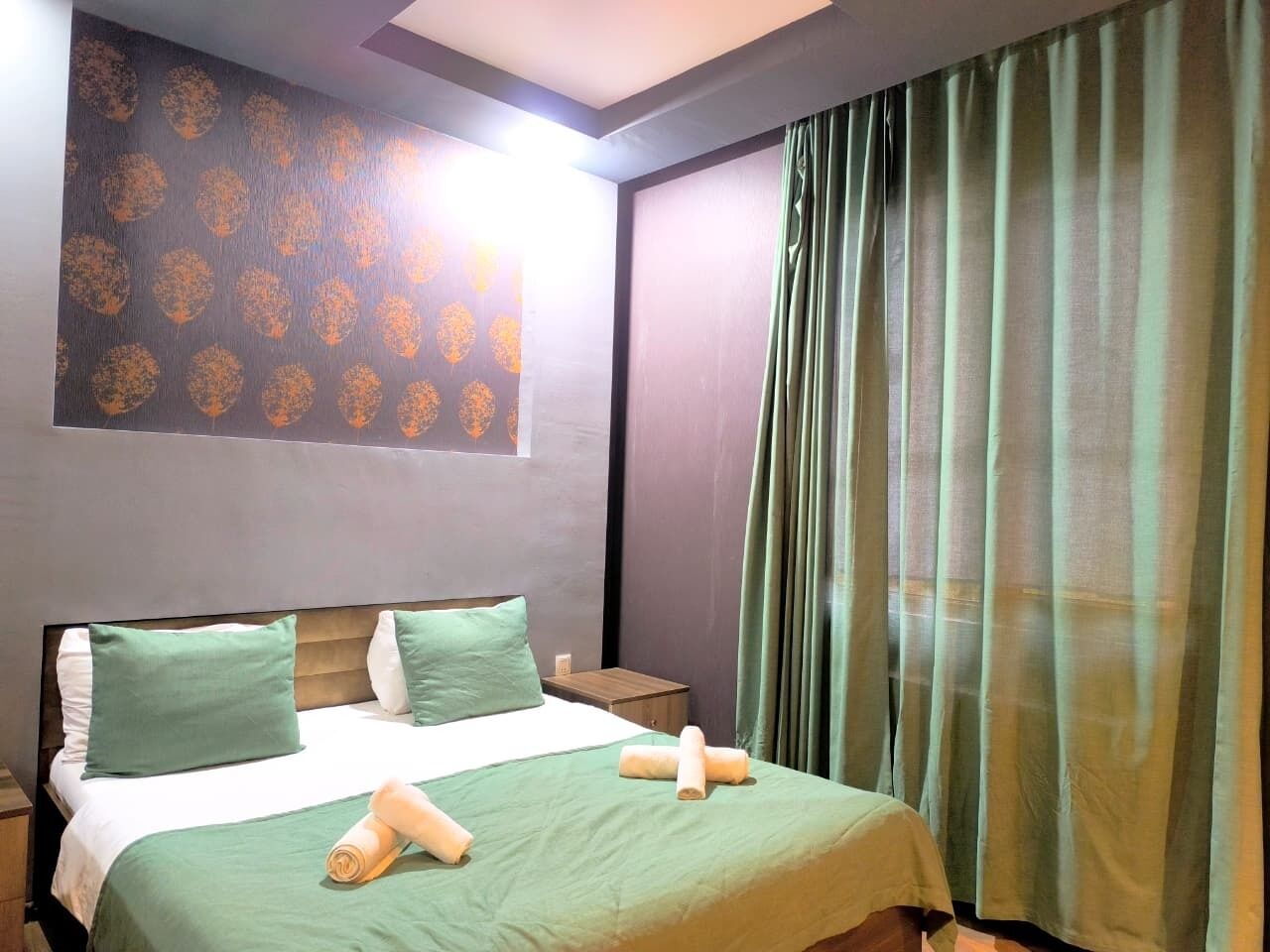 A hotel room with a bed, green pillows, a wooden headboard, a nightstand, and a window with curtains.